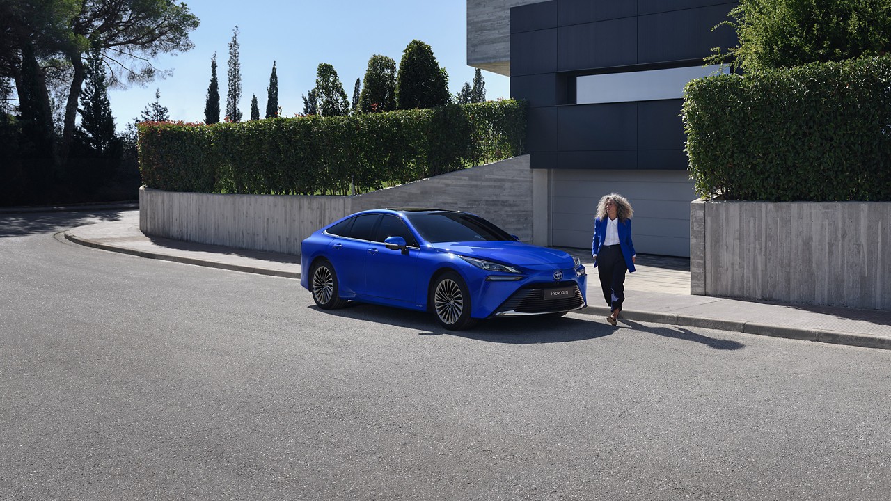 Nő kilép egy modern házból, és körbesétál egy élénk kék Toyota Mirai körül, amely a ház előtt parkol az utcán.